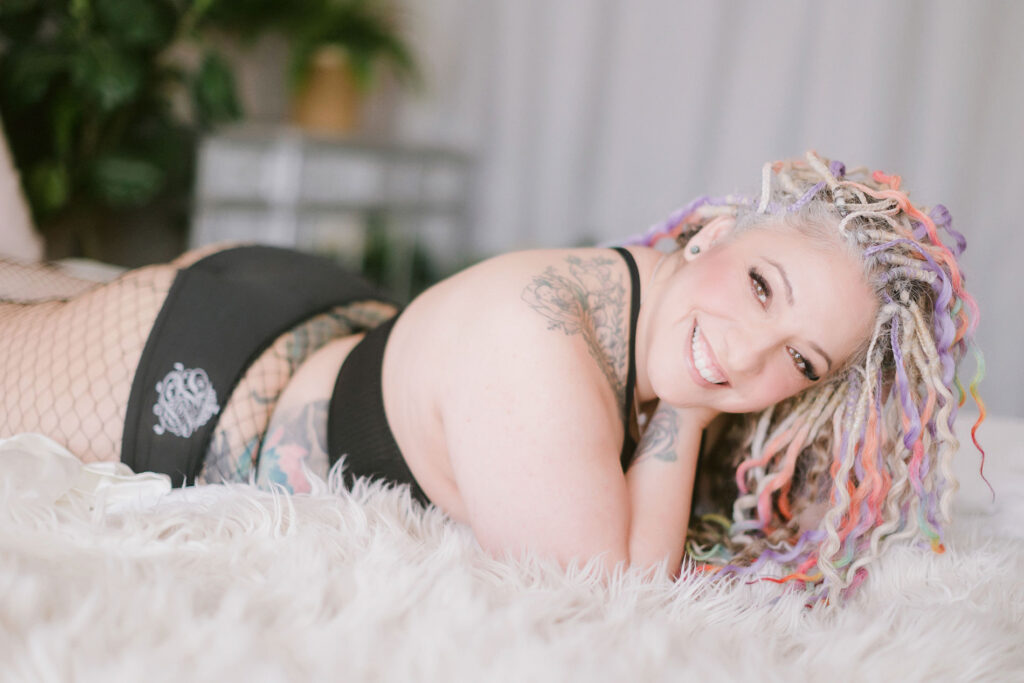 Playful boudoir portrait of a smiling woman with colorful locs, tattoos, and fishnet lingerie, lying on a soft white faux fur blanket. Captured at Heather Elizabeth’s Northern California boudoir studio, celebrating bold beauty, confidence, and body positivity.