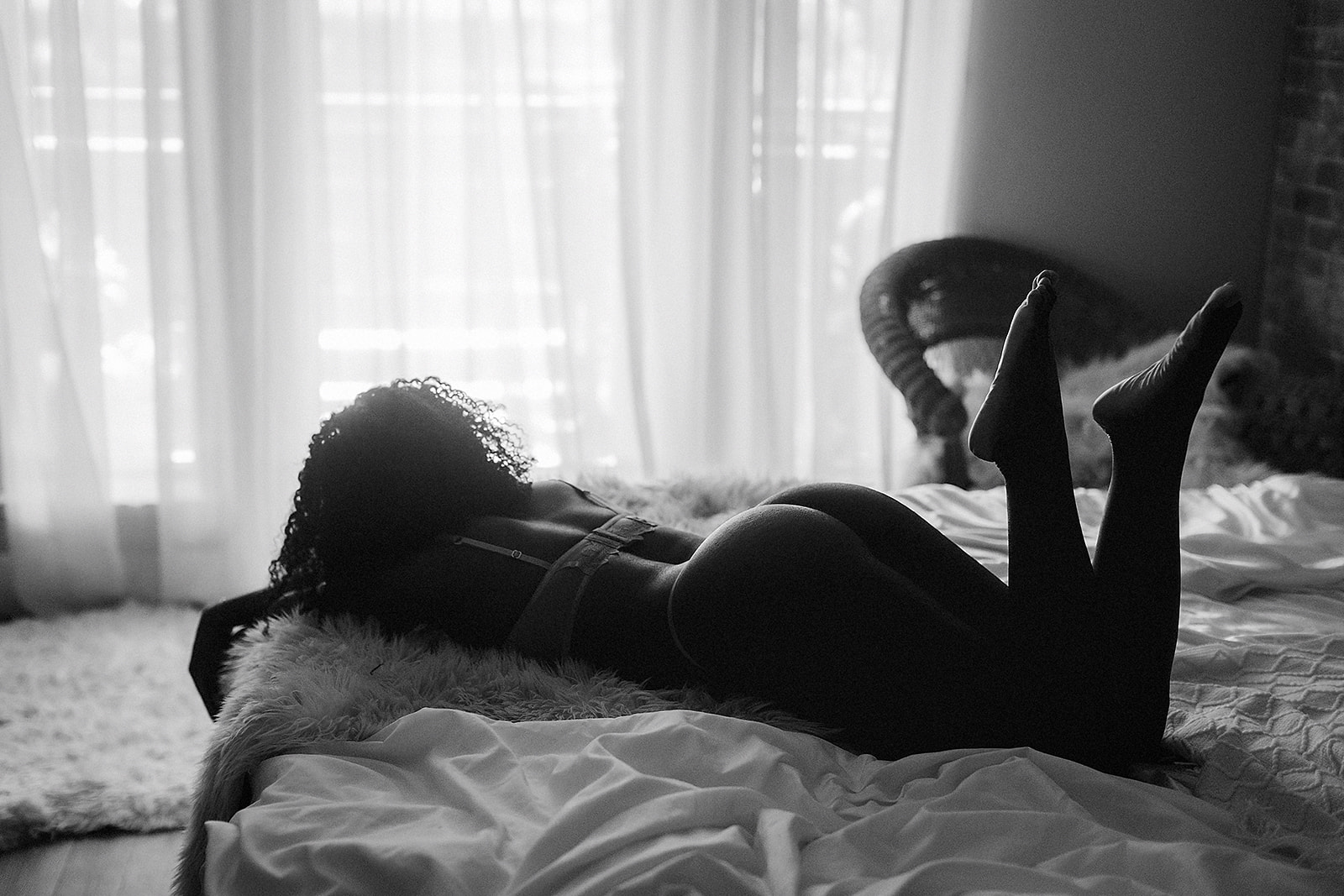 Black and white boudoir portrait of a woman laying on a bed in lingerie, backlit by window light, photographed by Northern California boudoir photographer Heather Elizabeth