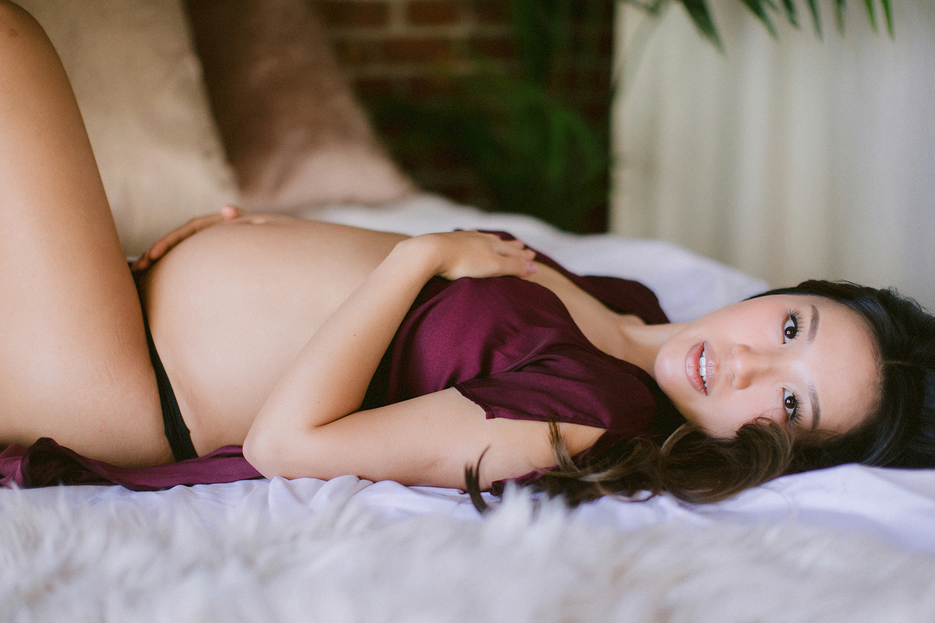 Pregnant woman reclining on a bed in a burgundy robe during a Sacramento maternity boudoir photography session