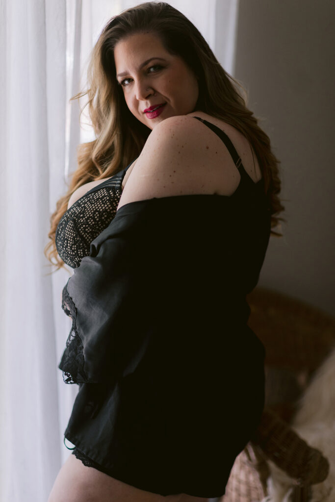 Woman in black lingerie and robe posing by a window during a Sacramento 40 Over 40 boudoir session